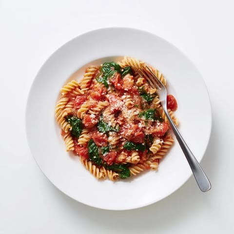 A close-up view of glossy rotini pasta coated in a savory tomato broth, flecked with wilted spinach leaves and melted Parmesan cheese.  