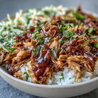 In a shallow bowl, fluffy white rice is generously topped with tender, shredded pulled pork. A colorful scoop of tangy coleslaw and a drizzle of smoky barbecue sauce complete this hearty Pulled Pork Bowl.